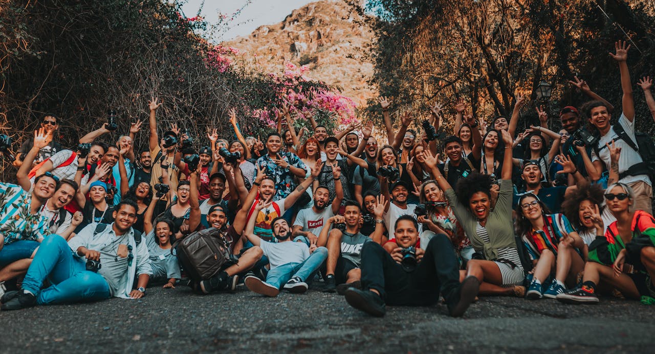 Group of multiracial happy people sitting with raised hands on asphalt road near mountains and trees in summer for portrait photo and looking at camera